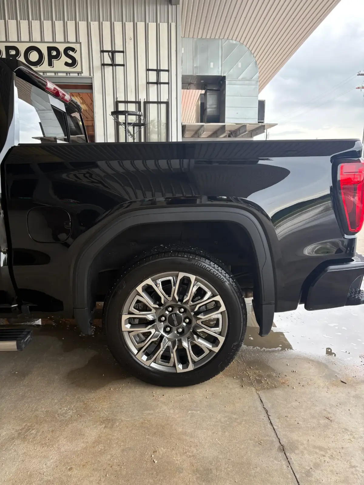 Rear side of a glossy black GMC pickup, showing its reflective bodywork, rear wheel and red taillight beside a metal-sided building. Water and suds are visible on the concrete.