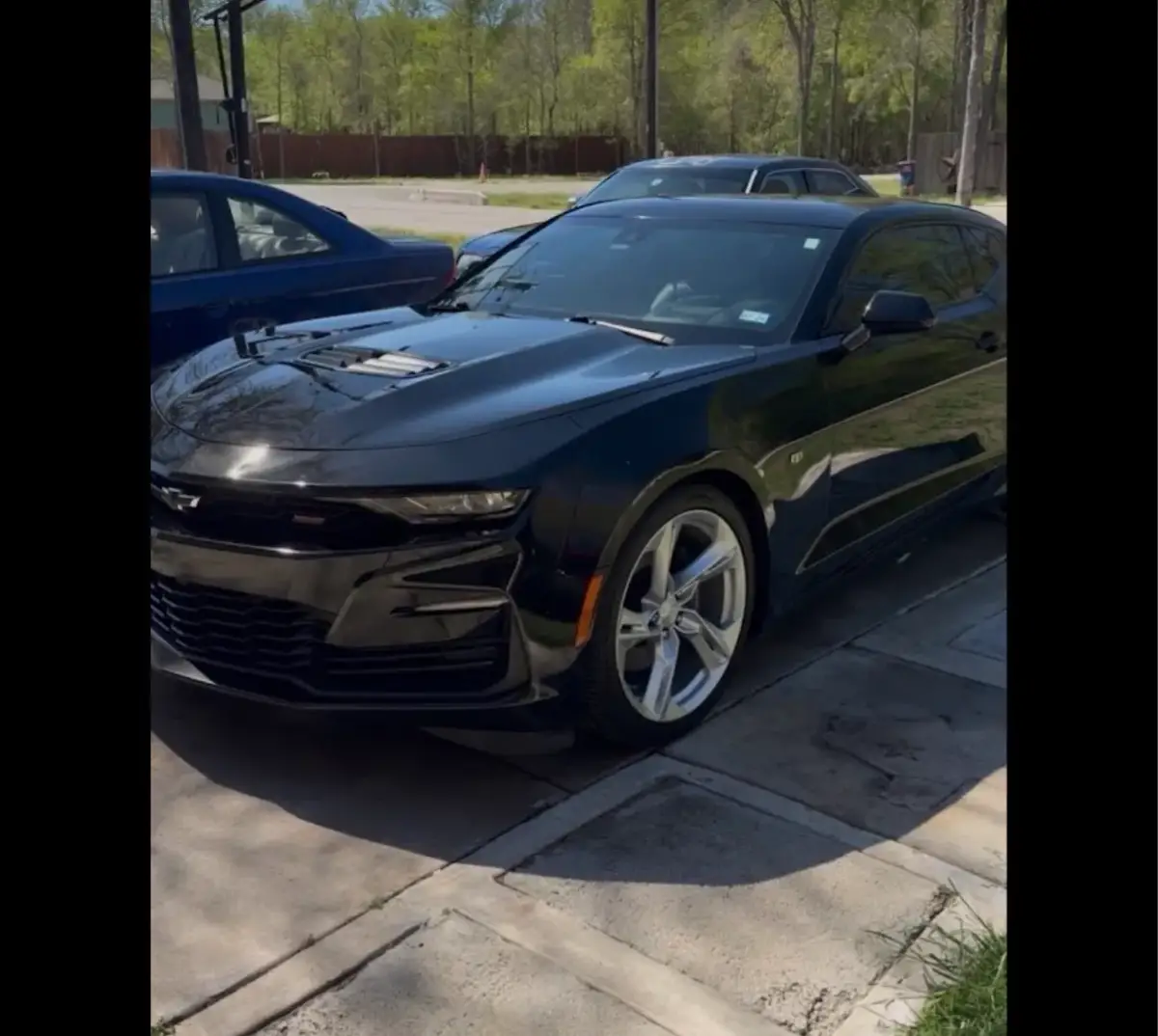 A shiny black Chevrolet Camaro parked on concrete, viewed from the front corner, with other cars and trees behind it. Black borders run along both sides of the image.