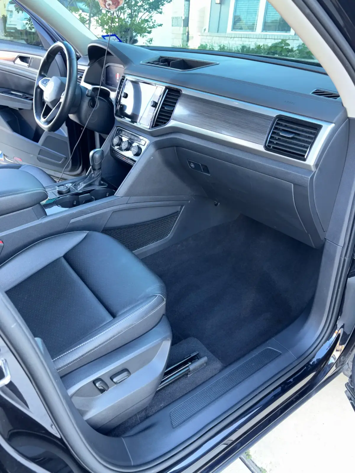 Car interior viewed through the open passenger door, showing a dark upholstered seat, carpeted footwell, dashboard, steering wheel and center console. No people or tools are visible.