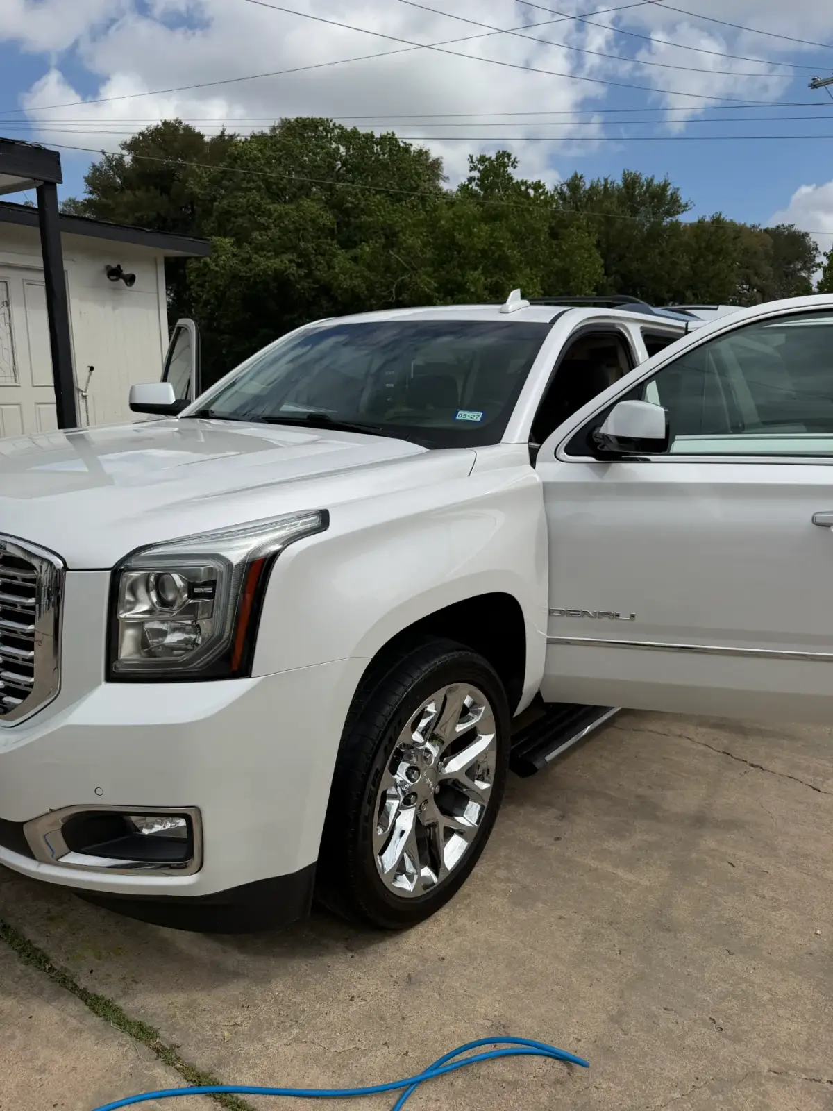 White Denali SUV parked on a concrete driveway with its front doors open. A blue hose lies in the foreground beside a white building.