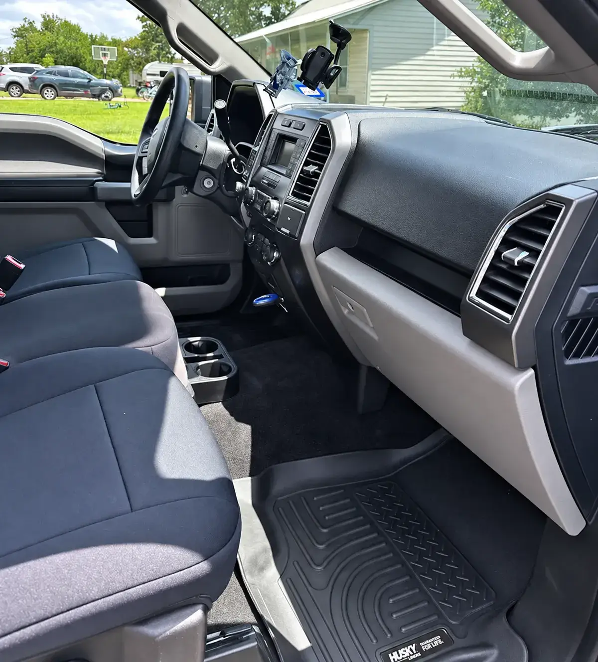 View into a truck’s front cabin, showing dark cloth seats, the steering wheel, dashboard, center controls, and a rubber passenger-side floor mat.