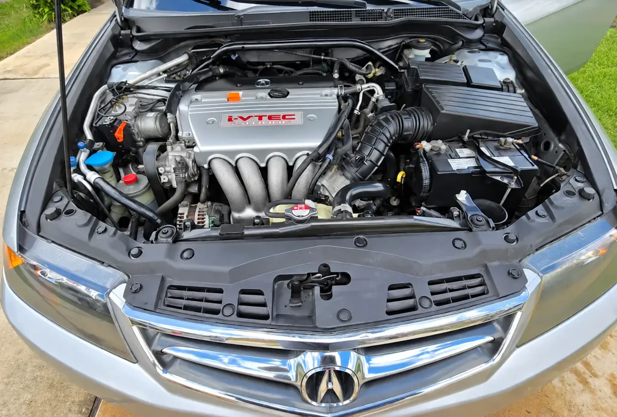 Open engine bay of a silver Acura parked outdoors, showing an i-VTEC engine, battery, hoses, fluid reservoirs, and front grille.
