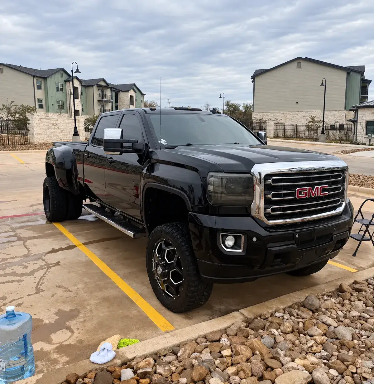 A black GMC pickup truck parked on wet pavement outside apartment buildings, with rocks and cleaning supplies in the foreground.