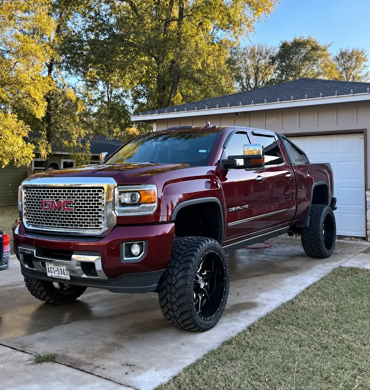 A burgundy GMC pickup with large off-road tires is parked on a concrete driveway in front of a residential garage, with trees overhead.